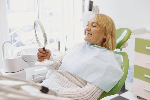 Smiling elderly patient sitting in a dental chair holding a mirror during a full mouth tooth restoration dental consultation at a modern dental clinic.