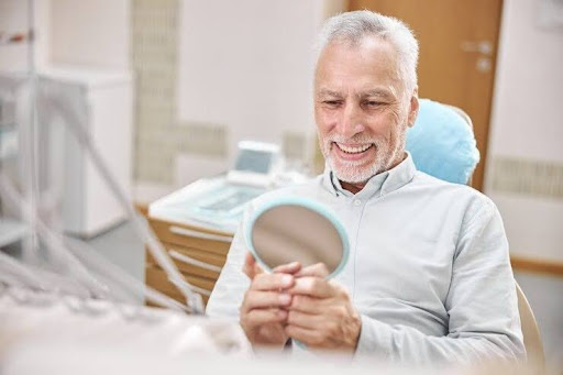 Happy senior patient smiling while holding a mirror during his free full mouth rehabilitation consultation.